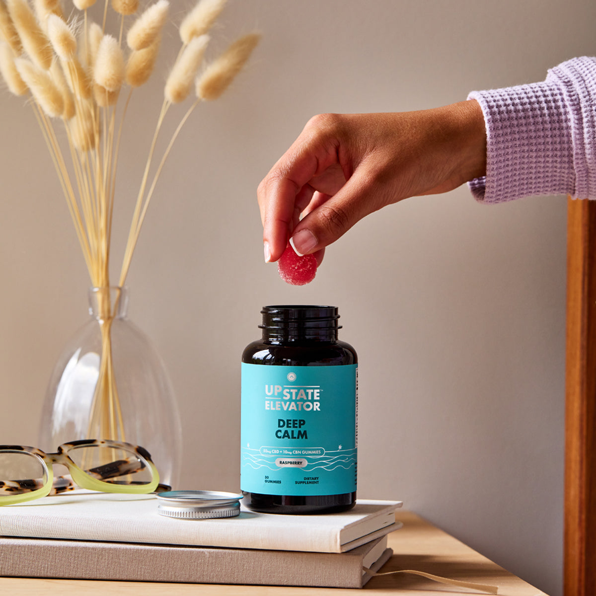 Hand holding a raspberry gummy above an open bottle of Upstate Elevator Deep Calm CBD gummies on a nightstand